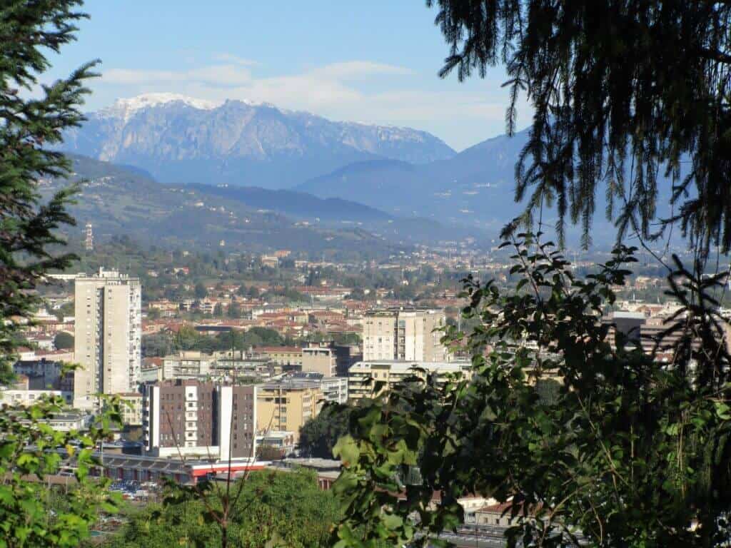 A scenic view of Vicenza, Italy, showcasing the contrast between the city's urban architecture and the lush greenery in the foreground, with the snow-capped peaks of the Dolomites looming in the background under a clear blue sky.