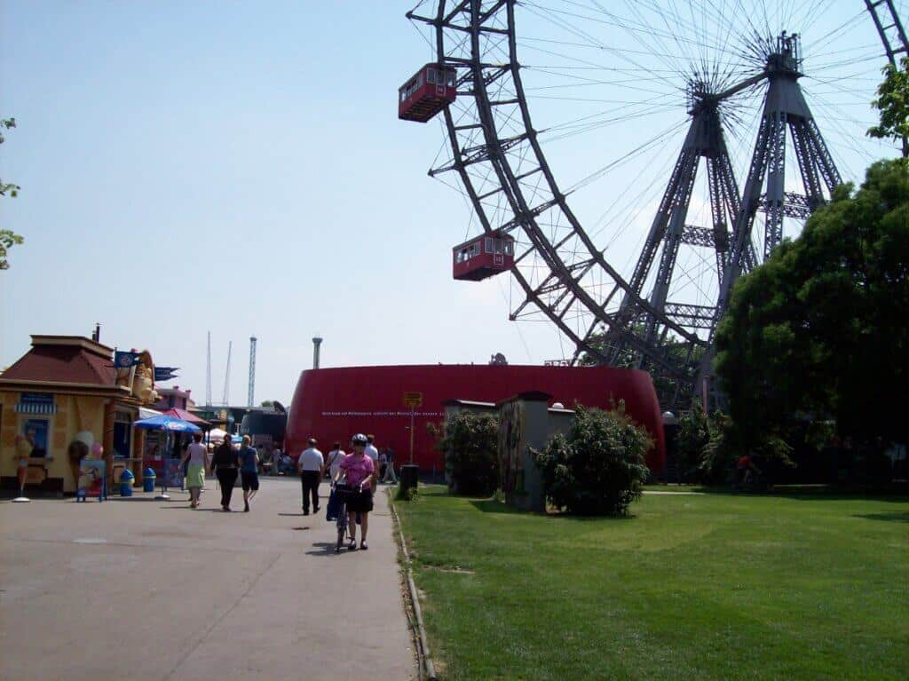 Visitors stroll along a wide walkway at the Prater amusement park in Vienna, with the iconic Wiener Riesenrad Ferris wheel towering in the background. The scene captures a clear day with vibrant greenery and various small vendor stalls and amusement park buildings along the path.