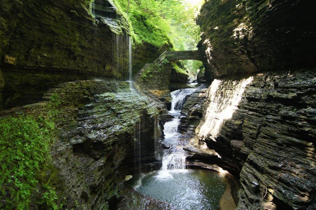 A serene view inside Watkins Glen State Park, showing a stone walkway that crosses over a cascading waterfall amidst deeply layered and dark shale rock formations. Sunlight filters through the lush greenery above, illuminating the misty water and creating a magical and tranquil atmosphere in this natural gorge.