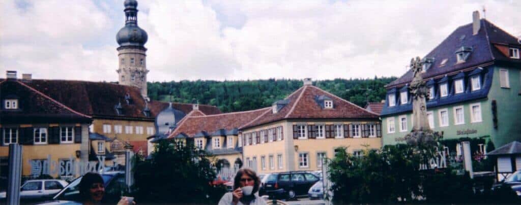 A snapshot of daily life in Weikersheim, Germany, featuring a public square surrounded by traditional, colorful buildings. The scene includes two people seated at a table, a statue prominently displayed in the center, and a historic building with a distinctive tower in the background. The lush green hills in the distance and parked cars suggest this is a blend of cultural heritage and modern lifestyle in a small town.