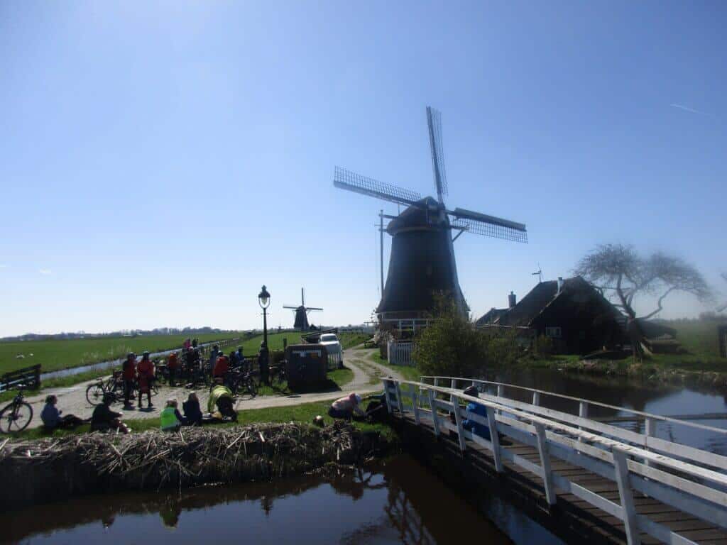 Cyclists rest and socialize near iconic Dutch windmills under a clear blue sky, with a tranquil canal in the foreground. The image captures a traditional scene in the Netherlands