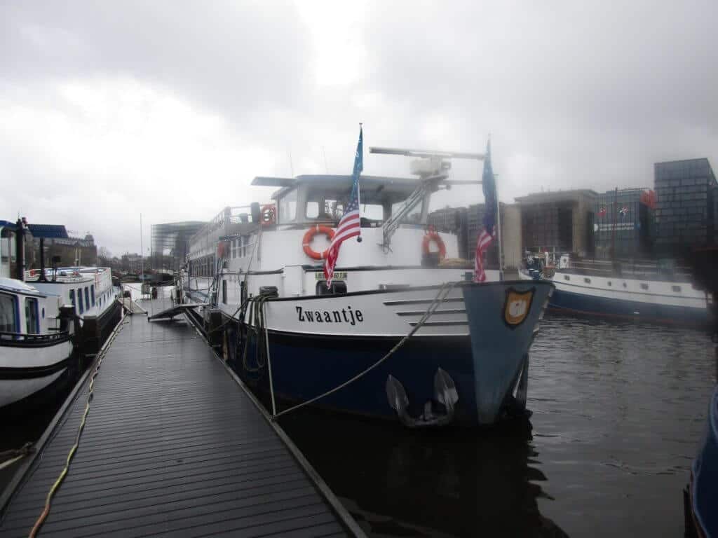 The 'Zwaantje' barge is moored in the misty Amsterdam harbor, with other canal boats and city buildings in the backdrop. The overcast sky sets a calm, muted tone over the scene, typical of a quiet day in the bustling maritime heart of Amsterdam.